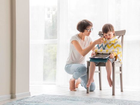 Mother cuts her son's hair by herself. Little boy sits, covered with cloth, and holds digital tablet. New normal in case of coronavirus COVID-19 quarantine and lockdown.