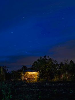 Star tracks over garden and gazebo illuminated with light bulbs. Long exposure of starry night sky.