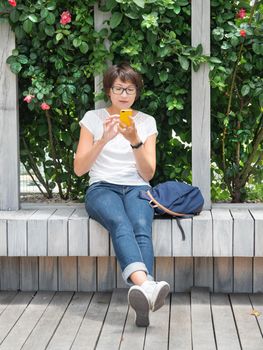 Woman is texting on smartphone. Pretty woman is sitting with smartphone on wooden bench in public park. Summer vibes.