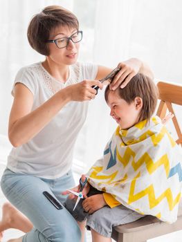 Mother cuts her son's hair by herself. Little boy sits, covered with cloth, and holds pair of scissors. New normal in case of coronavirus COVID-19 quarantine. Beauty at home lifestyle.