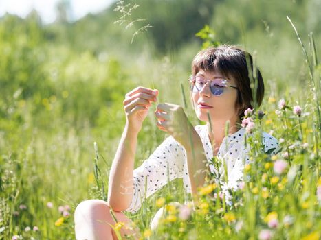 Woman in colorful sunglasses, enjoys sunlight and flower fragrance on grass field. Summer vibes. Relax outdoors. Self-soothing.