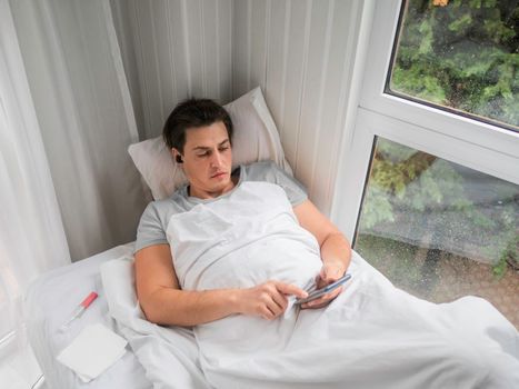 Sick man is lying in bed with thermometer. Man is searching online for disease symptoms or texting to his doctor. Using wireless technology in case of temporary disability.