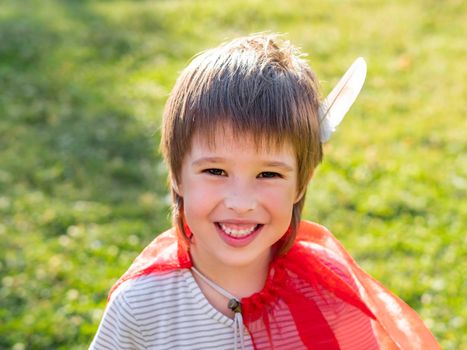 Portrait of smiling boy playing American Indian. Kid with white bird feather and red cloak. Costume role play. Outdoor leisure activity.