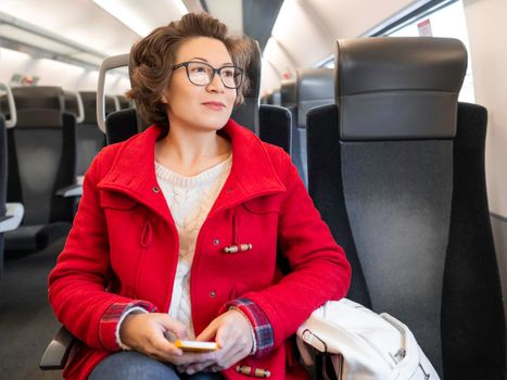 Smiling woman in red duffle coat with on smartphone sits near window in suburban train. Travel by land vehicle.