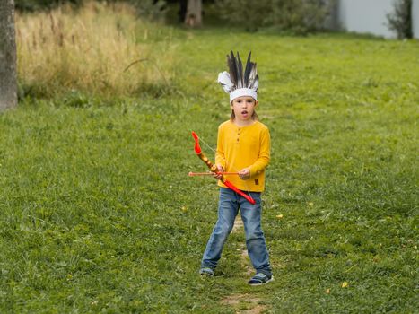 Little boy is playing American Indian on field. Kid has handmade headdress made of feathers and bow with arrows. Costume role play. Outdoor leisure activity. Fall season.