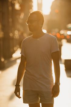 A man in sunglasses, a white t-shirt, and shorts is standing in the rays of the sunset on an old street in Spain. A guy with a beard is posing in Valencia in the evening.