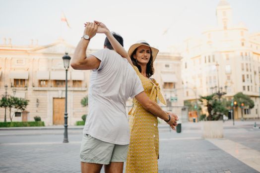 A happy girl in a hat and a yellow dress with a plunging neckline is dancing with her boyfriend with a beard and sunglasses in the old town. A couple of tourists on the sunset in Valencia.