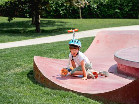 Little boy fell off kick scooter while riding in skate park. Special concrete bowl structures in urban park. Training to skate at summer.