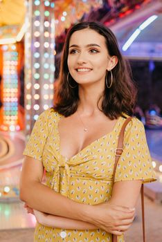 A young brunette woman in a yellow dress with a plunging neckline is staying between amusement rides. A girl at the fair in Valencia in the evening.