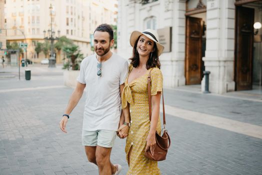 A happy girl in a hat and a yellow dress with a plunging neckline and her boyfriend with a beard and sunglasses are walking holding each other's hand in the old town. A couple of tourists in Valencia.