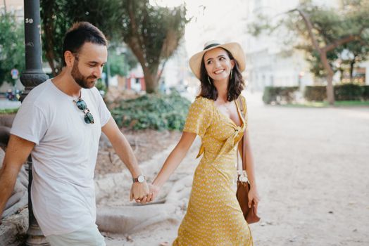 A smiling girl in a hat and a yellow dress and her boyfriend with a beard are walking holding each other's hand in a park in Spain. A happy couple of lovers are enjoying Valencia in the evening.