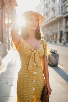 A pretty young woman in a yellow dress with a plunging neckline is holding a hat on her head on the sunset in Spain. A gorgeous smiling girl is standing on an old street in Valencia in the evening.