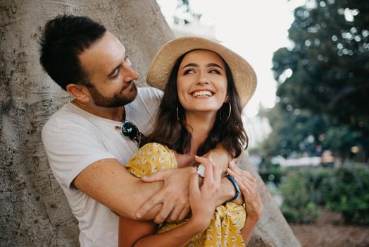 A smiling woman in a hat and a yellow dress and her happy boyfriend with beard are hugging under an old Valencian Ficus Macrophylla in Spain. A couple of lovers are enjoying Valencia in the evening.