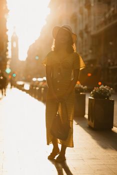 A cute young woman in a hat and a yellow dress with a plunging neckline is holding a leather bag on the sunset in Spain. A gorgeous smiling girl is posing on an old street in Valencia in the evening.