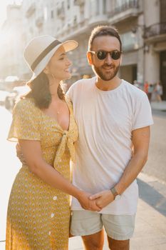 A girl in a hat and a yellow dress with a plunging neckline and her boyfriend with a beard are walking holding each other's hand in Spain. Couple of lovers hugging each other on the sunset in Valencia