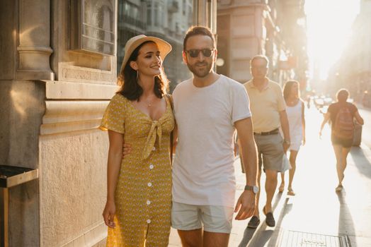 A man with a beard and sunglasses is hugging around the waist with his hand of a girlfriend in a hat and a yellow dress with a plunging neckline in Spain. A couple of lovers on the sunset in Valencia.