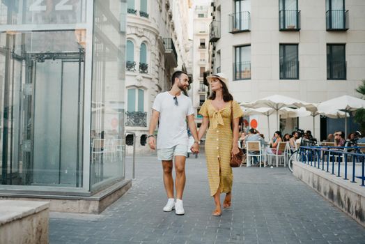 A happy girl in a hat and a yellow dress with a plunging neckline and her boyfriend with a beard and sunglasses are walking through the old town. A couple of tourists in the evening in Valencia.