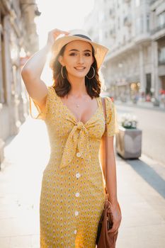 A cute young woman in a yellow dress with a plunging neckline is holding a hat on her head on the sunset in Spain. A gorgeous smiling girl is posing on an old street in Valencia in the evening.