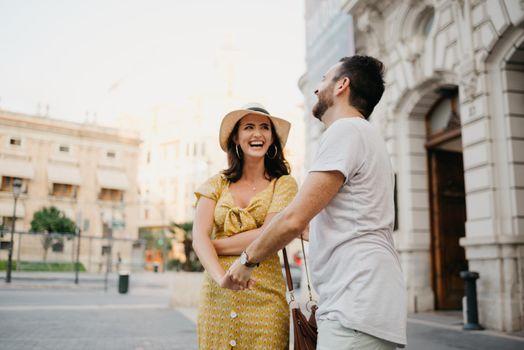 A laughing girl in a hat and a yellow dress with a plunging neckline is dancing with her boyfriend with a beard and sunglasses in the old town. A couple of tourists on the sunset in Valencia.