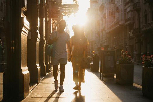 A girl in a hat and a yellow dress with a plunging neckline and her boyfriend with a beard are walking holding each other's hand in Spain. A couple of tourists on a date on the sunset in Valencia.