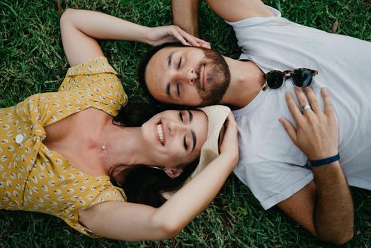 A brunette girl in a dress and a hat is lying near her boyfriend with a beard on the grass in the park in Valencia. A couple of tourists with closed eyes is enjoying the summer in Spain.