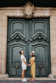 A girl in a hat and a dress with a plunging neckline and her boyfriend with a beard are staying near the giant doors holding each other's hand in the old town. A couple of tourists in Valencia.