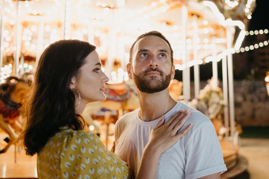 A close portrait of a young woman in a yellow dress and her boyfriend with a beard who are staying near the carousel. A couple of lovers on a date at the fair in Valencia.