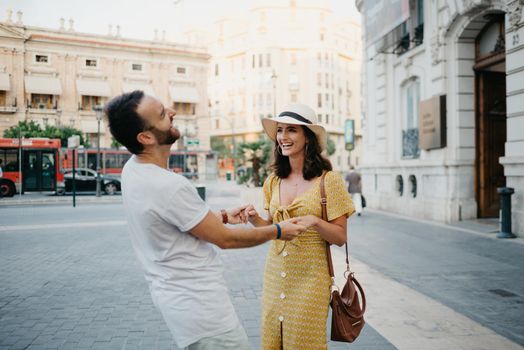A happy girl in a hat and a yellow dress with a plunging neckline is dancing with a boyfriend with a beard and sunglasses in the old town. A couple of tourists on the sunset in Valencia.