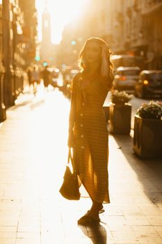 A cute young woman in a yellow dress with a plunging neckline is holding a leather bag on the sunset in Spain. A gorgeous smiling girl is posing on an old street in Valencia in the evening.
