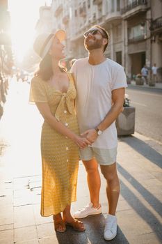 Girl in a hat and a yellow dress with a plunging neckline and her boyfriend with a beard are laughing holding each other's hand in Spain. Couple of lovers hugging each other on the sunset in Valencia