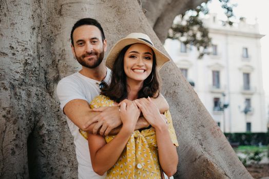A smiling girl in a hat and a yellow dress and her happy boyfriend with beard are hugging under an old Valencian Ficus Macrophylla in Spain. A couple of lovers are enjoying Valencia in the evening.