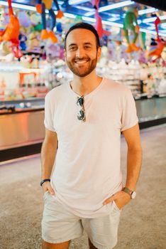 A stylish man with a beard and a sunglasses in the white t-shirt and shorts is staying between amusement rides. A guy at the fair in Valencia in the evening.