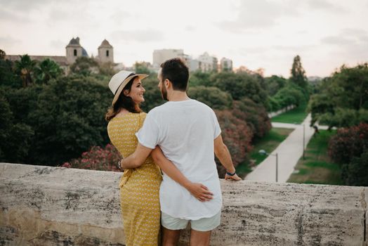 A girl in a yellow dress and a hat and her boyfriend is hugging each other on the ancient bridge in old Spain town. A couple of tourists is looking at each other in the park in the evening in Valencia