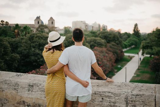 A girl in a yellow dress and a hat and her boyfriend is hugging each other on the ancient bridge in old Spain town. A couple of tourists is enjoying the view of the park in the evening in Valencia.