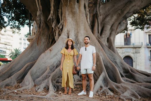 A beautiful young brunette in a hat and yellow dress with her boyfriend with a beard under an old giant Valencian Ficus Macrophylla tree in Spain in the evening. A couple of tourists in Valencia.