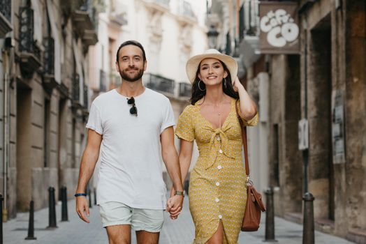 A girl in a yellow dress with a plunging neckline and her boyfriend with a beard are walking in old Spain town. A couple of tourists on a date in the evening Valencia. A lady is checking her hat.