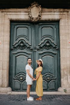 A happy girl in a hat and a dress with a plunging neckline and her boyfriend with a beard are posing near the giant doors holding each other's hand in the old town. A couple of tourists in Valencia.