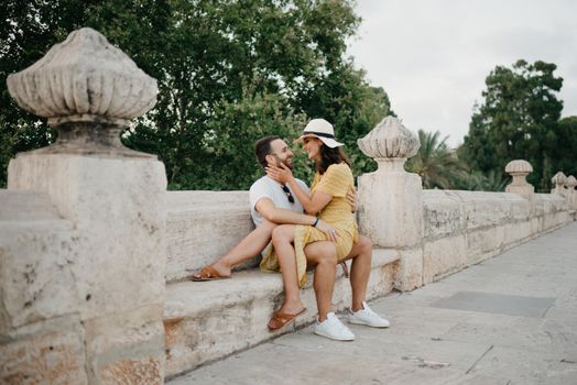 A girl in a hat and a yellow dress with a plunging neckline is sitting on the legs of her boyfriend with a beard on the ancient bridge in old Spain town. A couple of tourists on a date in Valencia.