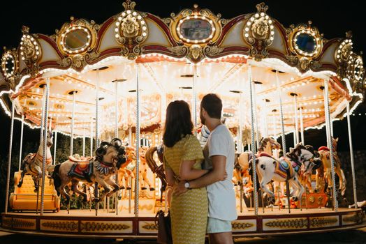A young woman in a yellow dress and her boyfriend with their backs are staring at the carousel. A couple of lovers on a date at the fair in Valencia.