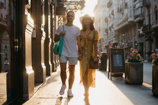 A girl in a hat and a yellow dress with a plunging neckline and her boyfriend with a beard and sunglasses are walking holding each other's hand in Spain. A couple of tourists on the sunset in Valencia