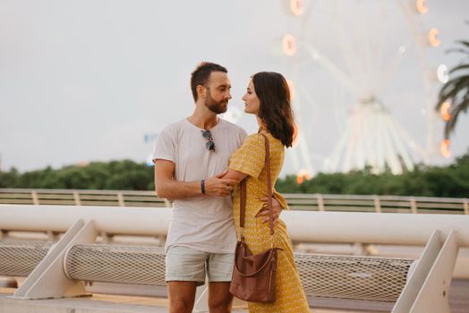 A brunette girl in a yellow dress and her boyfriend are hugging on a bridge with a Ferris wheel in the background in Valencia. A couple of tourists on a date in the warm evening.