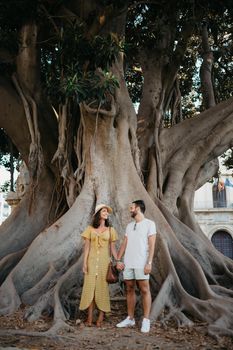 A beautiful young brunette in a hat and yellow dress with her boyfriend with a beard under an old giant Valencian Ficus Macrophylla tree in Spain in the evening. A couple of tourists enjoying Valencia