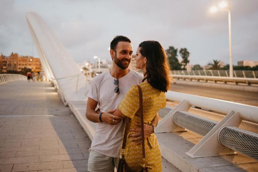 A brunette girl in a yellow dress and her smiling boyfriend are hugging on a white bridge in Valencia. A couple of tourists on a date in the warm evening.