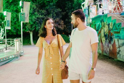 A woman in a yellow dress with a plunging neckline and a man with a beard are walking holding each other's hands between amusement rides. A couple of lovers on a date at the fair in Valencia.