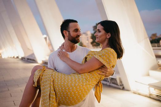 A man with a beard is holding his girl in a yellow dress in his arms on a white bridge in Valencia. A couple of tourists on a date in the warm evening.