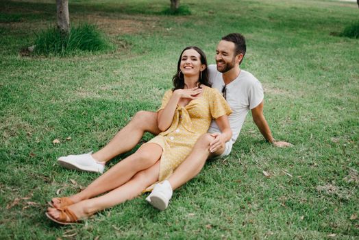 A girl in a yellow dress is sitting between the legs of her boyfriend on the grass.