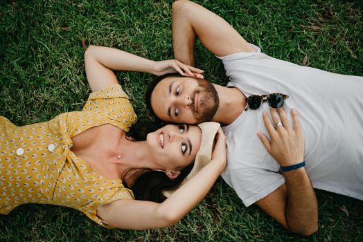 A brunette girl in a dress and a hat is lying near her boyfriend with a beard on the grass in the park in Valencia. A couple of tourists is enjoying the summer in Spain.