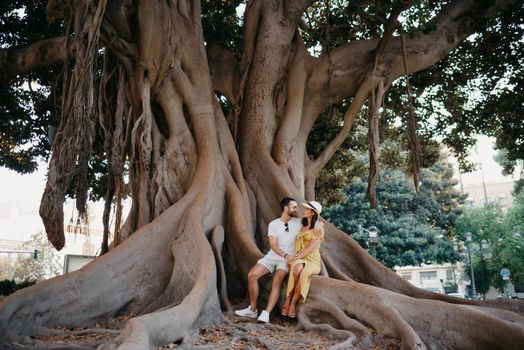 A young woman in a hat and a yellow dress and her boyfriend with a beard hug sitting on a root of an old Valencian Ficus Macrophylla in Spain. A couple of tourists are enjoying Valencia in the evening