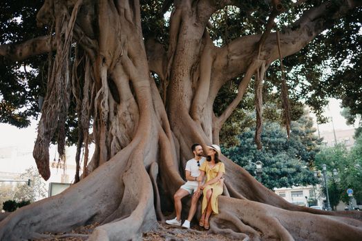 A young woman in a hat and a yellow dress sits near her boyfriend with a beard on a root of an old Valencian Ficus Macrophylla tree in Spain. A couple of tourists are exploring Valencia in the evening