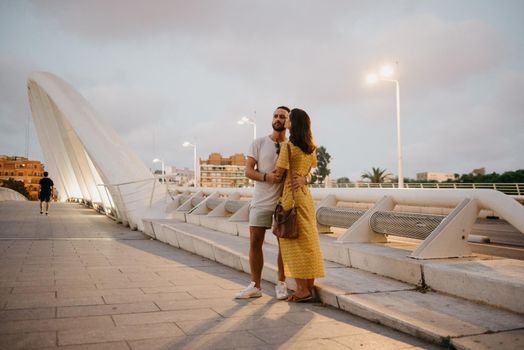 A brunette girl in a yellow dress and her smiling boyfriend are hugging on a white bridge in Valencia. A couple of tourists in a full length on a date in the warm evening.